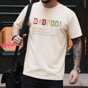 May include: A cream-colored t-shirt with the text "DADPOOL" in red, purple, green, and pink. The shirt includes a definition: "like a dad, only cooler." The shirt also says "See also: handsome, asshole."