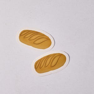 Könnte beinhalten: Zwei Aufkleber mit einem goldbraunen Brotlaib mit detaillierten Linien. Jeder Brot-Aufkleber hat einen weißen Rand, vor einem weißen Hintergrund.