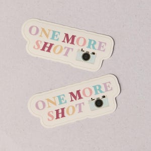 May include: Two stickers with the text "ONE MORE SHOT" in colorful letters. Each sticker has a light blue camera illustration. The stickers are on a light gray background.