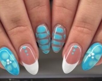Pode incluir: Unhas ovais com um design azul claro e branco. Algumas unhas apresentam uma ponta francesa branca, enquanto outras têm um padrão floral azul e branco. O design inclui uma base transparente com listras azuis.