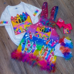 Puede incluir: Una camiseta blanca con un gráfico de caja de crayones coloridos y el nombre Kymbrelle 3, junto con un vestido de peto arcoíris con personajes de crayones, un dobladillo de plumas, un lazo para el pelo y calcetines con volantes arcoíris.