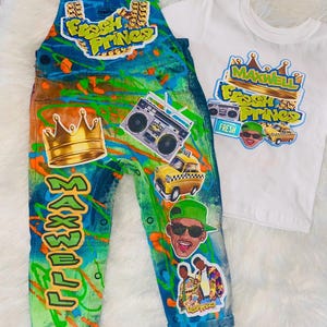 Puede incluir: Un conjunto de peto vaquero y camiseta blanca. El peto está decorado con pintura estilo grafiti, una corona dorada, un radiocasete e imágenes de un taxi y una persona con gafas de sol. La camiseta tiene un gráfico a juego.