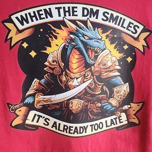 Op de afbeelding: Rood t-shirt met een gedetailleerde illustratie van een blauwe draak in gouden harnas, met een zwaard. De tekst "WHEN THE DM SMILES IT'S ALREADY TOO LATE" staat op een banner. De draak staat tegen een zwarte achtergrond met sterren en vlammen.