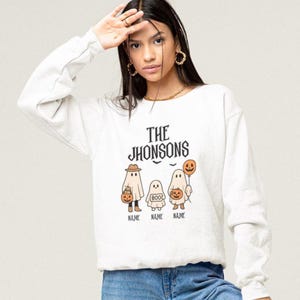 May include: A white long-sleeve sweatshirt featuring a graphic print with the text THE JHONSONS above three ghost figures. The ghosts are holding pumpkins and a balloon, with the word BOO on one ghost and the placeholder text NAME below each.