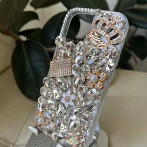 May include: Clear phone case bedazzled with numerous clear rhinestones, gold-toned floral accents, and a gold crown with the word "Juicy". The case is on a silver phone stand.