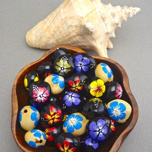 May include: A wooden bowl filled with hand-painted wooden beads. The beads are black with colourful floral designs. The bowl is sitting on a grey surface.