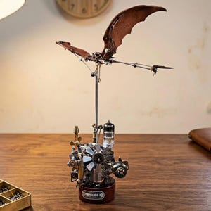 May include: A steampunk-style metal sculpture of a bat with outstretched wings. The wings are a dark brown color, and the body features intricate mechanical details. The sculpture is mounted on a dark red base and is displayed on a wooden surface.