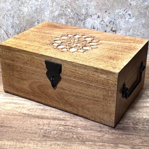 Könnte beinhalten: Eine rechteckige Holzkiste mit einem geschnitzten Blumendesign auf dem Deckel. Die Kiste ist hellbraun mit einem schwarzen Metallverschluss und Griff. Die Holzmaserung ist sichtbar, und die Kiste steht auf einer Holzoberfläche.