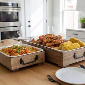 Puede incluir: Dos bandejas de madera con asas metálicas, cada una con comida. Una bandeja tiene paella, la otra maíz a la parrilla y brochetas. Las bandejas están sobre una mesa de madera con platos y cubiertos.