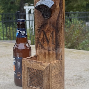 May include: Wooden bottle opener with a built-in cap catcher. The opener is mounted on a rectangular wooden stand with a decorative initial "M" and a metal bottle opener. A beer bottle is next to the opener.