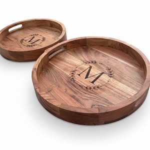 May include: Two wooden serving trays. One is oval, the other is round. Both have a dark wood grain and a raised edge. Each tray features a decorative wreath design with the letter "M" in the center. The oval tray has a handle cutout.