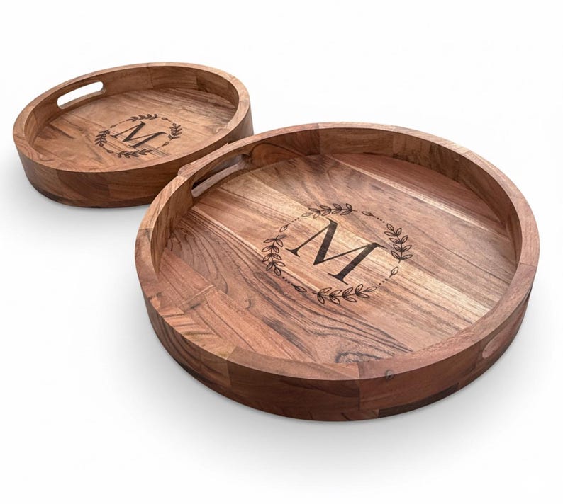 Puede incluir: Dos bandejas de madera para servir. Una es ovalada, la otra redonda. Ambas tienen una veta de madera oscura y un borde elevado. Cada bandeja presenta un dise&ntilde;o de corona decorativa con la letra "M" en el centro. La bandeja ovalada tiene un recorte para el asa.