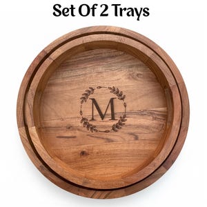 Puede incluir: Juego de 2 bandejas redondas de madera con borde elevado. Las bandejas presentan una inicial "M" grabada dentro de una corona de laurel. La veta de la madera es visible y las bandejas tienen un color marr&oacute;n c&aacute;lido. El texto "Set Of 2 Trays" est&aacute; en la parte superior.