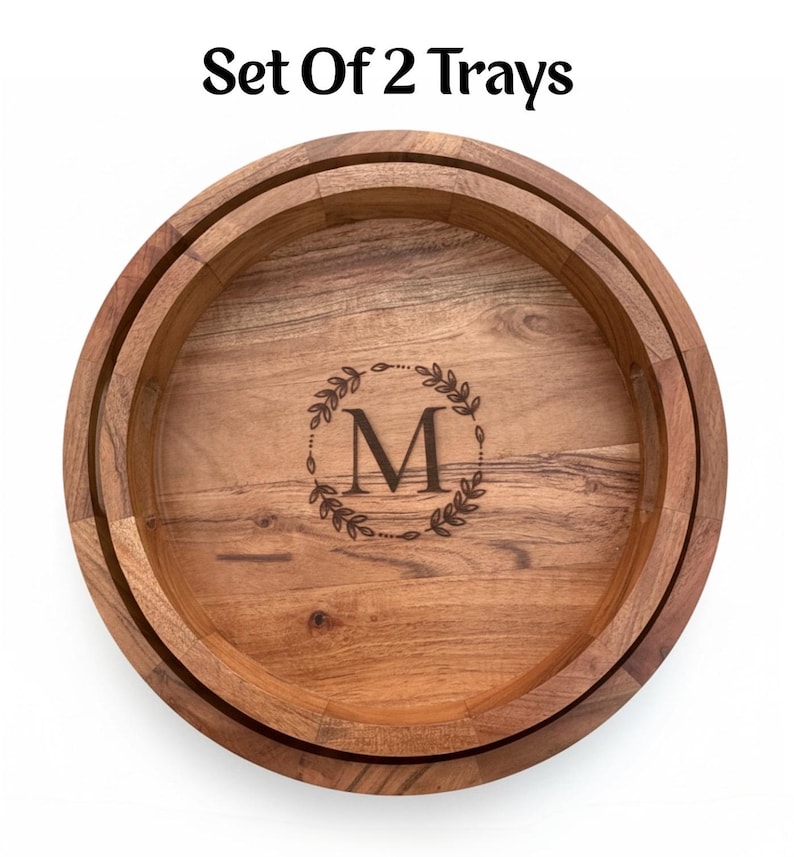 Puede incluir: Juego de 2 bandejas redondas de madera con borde elevado. Las bandejas presentan una inicial "M" grabada dentro de una corona de laurel. La veta de la madera es visible y las bandejas tienen un color marr&oacute;n c&aacute;lido. El texto "Set Of 2 Trays" est&aacute; en la parte superior.