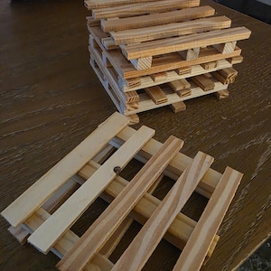 May include: A stack of light-colored wooden pallet coasters and one individual pallet coaster placed in front. Each coaster is constructed from thin, parallel wooden slats arranged in a grid pattern, showcasing the natural grain and light tan color.