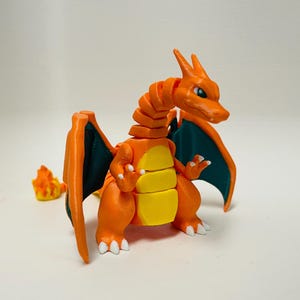 Könnte beinhalten: 3D-gedruckte Charizard-Figur mit orangefarbenen und gelben Körpersegmenten, dunkelgrünen Flügeln und weißen Krallen. Eine kleine Flamme befindet sich links. Ein Sammlerobjekt.