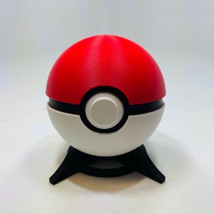 May include: A red and white Poké Ball replica, a spherical device with a black band and a central white button. The ball is set on a black, three-legged stand, and is a collectible item.