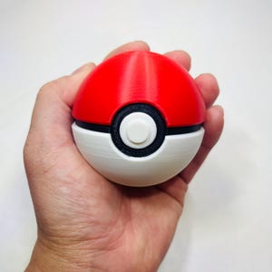 May include: A red and white Poké Ball with a black band and a white button in the center. The ball is held in a hand, showcasing its spherical shape and design. The top half is red, and the bottom half is white.