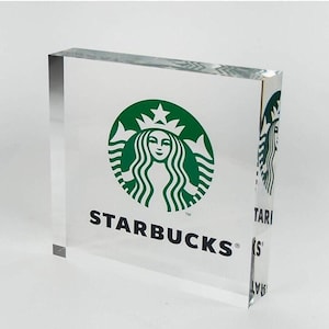 Puede incluir: Un bloque acrílico transparente con el logotipo de Starbucks. El logotipo es un emblema verde de sirena con el texto "STARBUCKS" en negro. El bloque es rectangular y se mantiene en posición vertical.