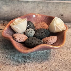 May include: A pink ceramic dish holds decorative items. The dish contains two white wooden roses, several porous lava stones in shades of pink, gray, and black, and a speckled gray stone. The dish has a wavy, organic shape.