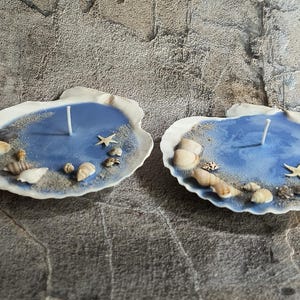 May include: Two seashell-shaped candles with a blue wax interior, resembling a small ocean. Each candle is decorated with sand, seashells, and a starfish. A white candle wick is centered in each blue wax pool.