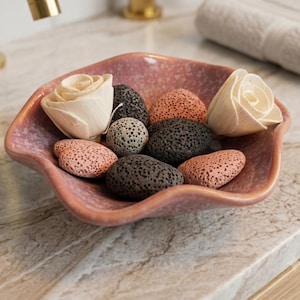 May include: A wavy-edged, speckled pink ceramic bowl containing two cream-colored wooden rose flowers, several dark gray porous lava stones, and three smooth, light pink porous stones arranged in a decorative cluster.
