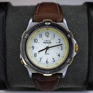 Pode incluir: Um relógio de pulso Timex Indiglo com mostrador branco, algarismos arábicos pretos, uma escala interna de 24 horas e um símbolo de resistência à água. O relógio possui uma caixa prateada, um bisel rotativo preto e uma bracelete de couro castanho.