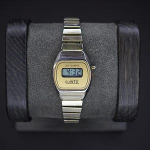 May include: A vintage digital watch with a silver band and a gold-toned face. The watch displays the time 4:32 on a small LCD screen. The brand name "QUINTEL" is printed below the screen. The watch is displayed on a gray surface.