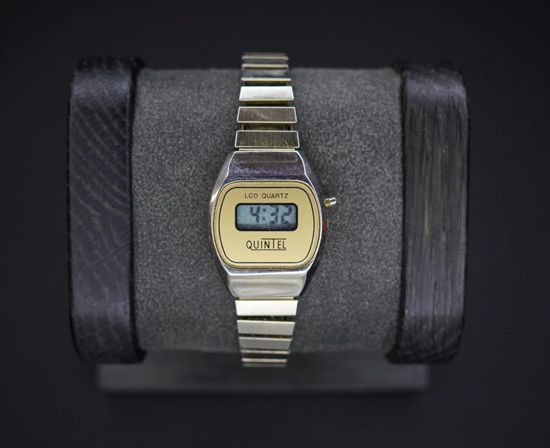 May include: A vintage digital watch with a silver band and a gold-toned face. The watch displays the time 4:32 on a small LCD screen. The brand name "QUINTEL" is printed below the screen. The watch is displayed on a gray surface.