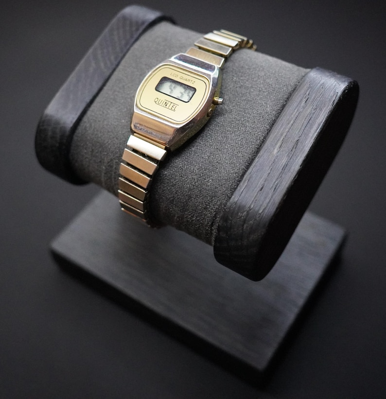 May include: A gold-toned Quintel digital watch with a rectangular face and a segmented metal band. The watch displays the time on a small LCD screen. The watch is displayed on a dark gray, padded watch stand with a black wooden base.