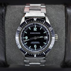May include: A silver-tone stainless steel wristwatch with a black dial featuring the text KINGSMARK, WATERPROOF, and SHOCKRESISTANT. The watch has a black rotating bezel with white markers, luminous hands, Arabic numerals at 6, 9, and 12, and a date window.