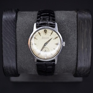 May include: A vintage Bulova watch with a silver-tone case and a black leather band. The watch face is cream-colored with black hands and hour markers. The watch is marked "Bulova Waterproof". The watch is displayed on a gray cushion.
