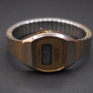 May include: A vintage gold-toned digital watch with a rectangular face and a silver-colored, expandable band. The watch face displays the text "LCD QUARTZ" and "QUINTEL". The watch is set against a dark background.