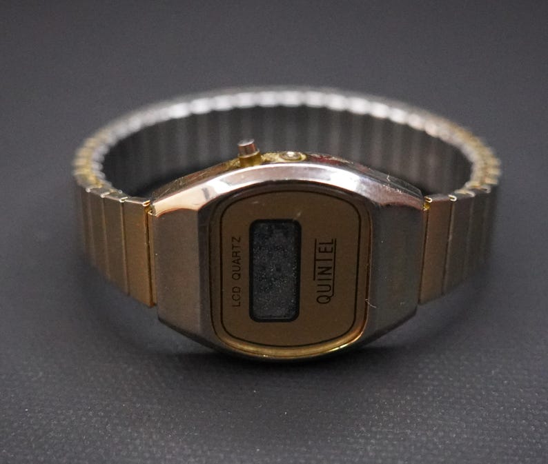 May include: A vintage gold-toned digital watch with a rectangular face and a silver-colored, expandable band. The watch face displays the text "LCD QUARTZ" and "QUINTEL". The watch is set against a dark background.