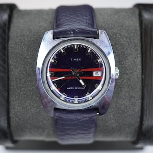 May include: A vintage Timex watch featuring a navy blue face with red stripes and a date display. The watch has a silver-tone case and a navy blue leather strap. The watch face displays the brand name "TIMEX" and the text "WATER RESISTANT".