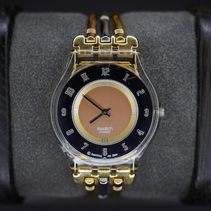 Op de afbeelding: Een Swatch polshorloge met een doorzichtige kunststof behuizing en een goudkleurige metalen band. De wijzerplaat heeft een zwarte buitenring met witte Arabische cijfers, een gouden binnencirkel en een effen beige centrum. Swatch Swiss AG 2004 is zichtbaar.