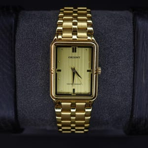 May include: Gold-coloured rectangular Orient watch with a matching link strap. The watch face is gold with black markers and hands. The text "ORIENT" and "WATER RESIST" are visible. The watch is displayed on a dark grey surface.