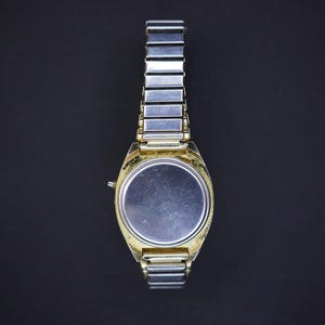 May include: A vintage wristwatch with a gold-toned case and a silver-toned metal band. The watch face is a plain silver color. The watch band is made of small, rectangular links. The watch is shown against a dark background.