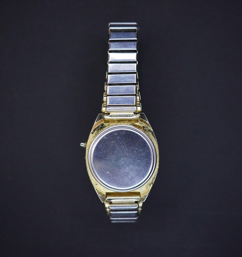 May include: A vintage wristwatch with a gold-toned case and a silver-toned metal band. The watch face is a plain silver color. The watch band is made of small, rectangular links. The watch is shown against a dark background.