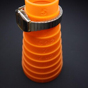May include: An orange watch sizing tool with measurements in inches, with a silver and gold watch resting on top. The tool is cylindrical and has measurements printed on the side. The background is black.