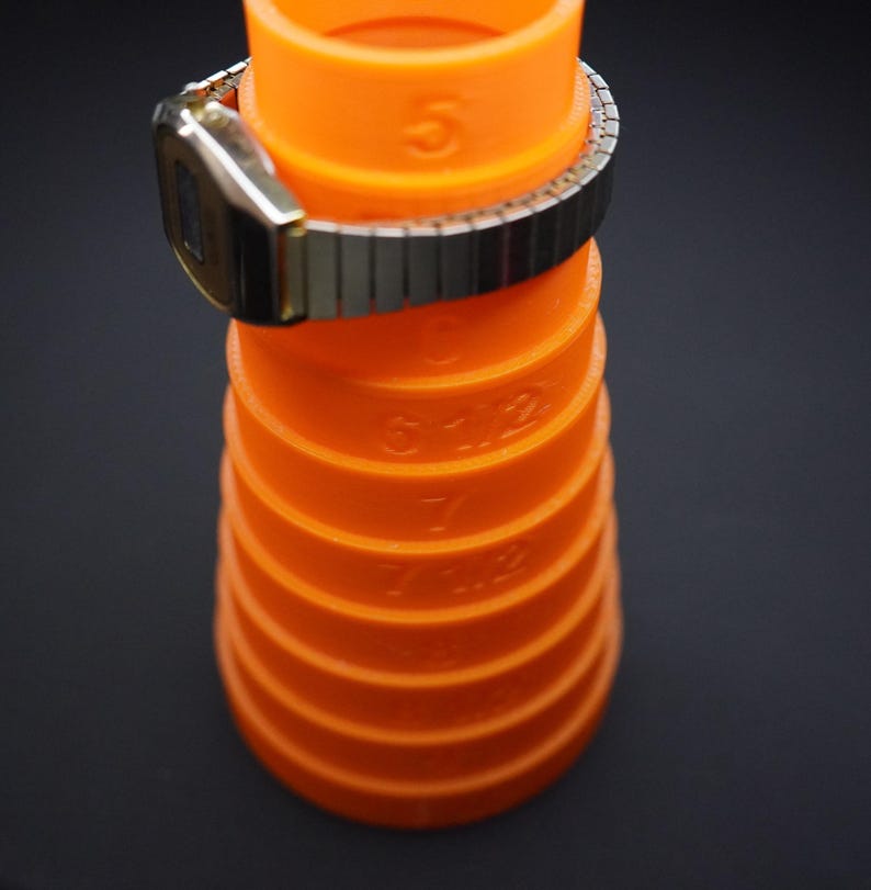 May include: An orange watch sizing tool with measurements in inches, with a silver and gold watch resting on top. The tool is cylindrical and has measurements printed on the side. The background is black.