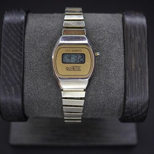 May include: A vintage silver-toned watch with a gold-colored face displaying the time 4:32. The watch has a rectangular LCD display and the brand name "QUINTEL" is printed below the time. The watch is on a gray watch stand.