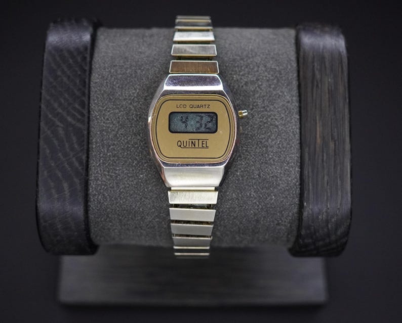 May include: A vintage silver-toned watch with a gold-colored face displaying the time 4:32. The watch has a rectangular LCD display and the brand name "QUINTEL" is printed below the time. The watch is on a gray watch stand.