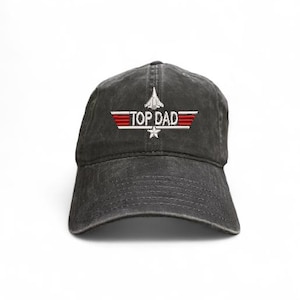 May include: A black baseball cap with the embroidered text "TOP DAD" in white, red, and silver. The design includes a stylized airplane and star. The cap has a curved brim and adjustable strap.