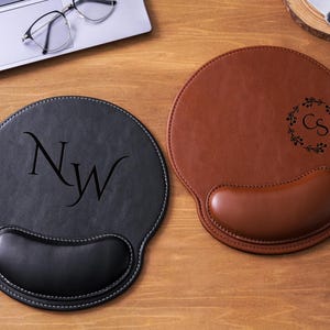 Op de afbeelding: Twee ronde leren muismatten met polssteunen. Eén is zwart met witte stiksels en de initialen "NW". De andere is bruin met een bloemenkrans en de initialen "CS". Beide liggen op een houten oppervlak.