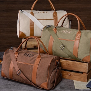 Op de afbeelding: Drie canvas duffeltassen in beige, olijfgroen en bruin, met bruine leren accenten. Elke tas heeft een handvat aan de bovenkant en een schouderriem. De tassen zijn gepersonaliseerd met geborduurde initialen. Een blauwe baseballpet staat op de voorgrond.