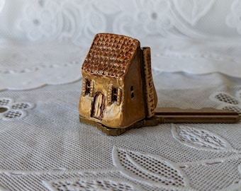 May include: A small, brown, ceramic house-shaped key holder with a gold-colored metal key attached.