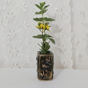 May include: A small, rectangular ceramic vase with a dark, mottled glaze and a carved floral motif on the front. The vase contains a single green stem with small, bright yellow flowers blooming near the top, creating a simple, natural display.