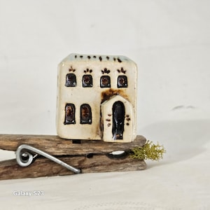 May include: A small, white ceramic house with brown accents and windows. The house is sitting on a wooden clothespin.