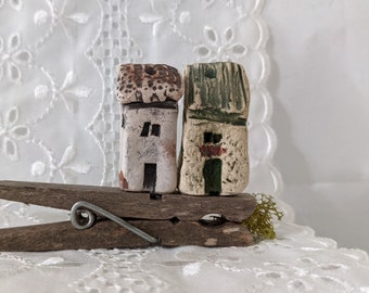 May include: Two small, hand-painted ceramic houses with brown, white, and green accents are perched on a wooden clothespin. The houses have small windows and doors.